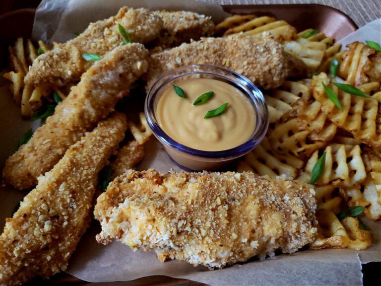 parchment lined mini cooking sheet with crispy chicken tenders, waffle fries and dipping sauce garnished with scallions