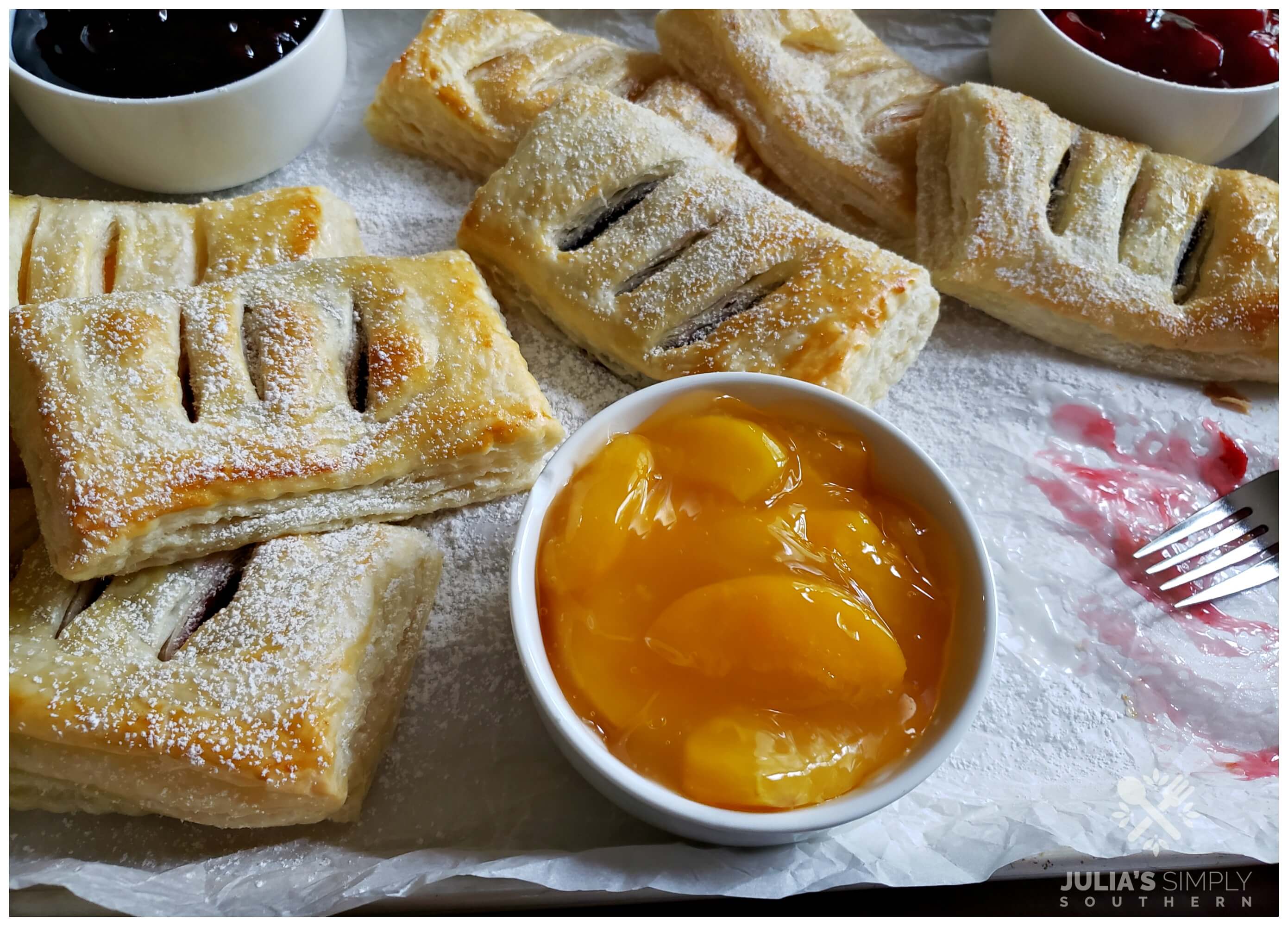 Assorted fruit hand pies on a confectioner's sugar coated sheet pan with extra fruit filling to be used for topping. Hand pies can be served alone or with a glazed topping or alongside a scoop of ice cream.