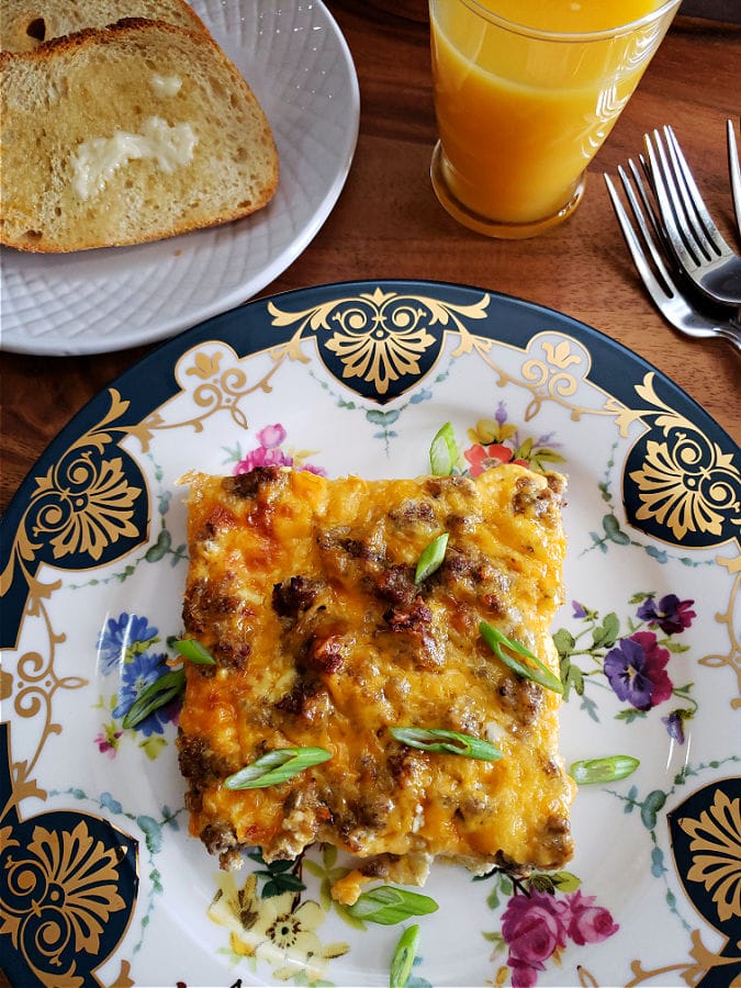 Breakfast casserole serving on a beautiful Biltmore Vanderbilt china plate served alongside a glass of orange juice and plate of toasted bread.