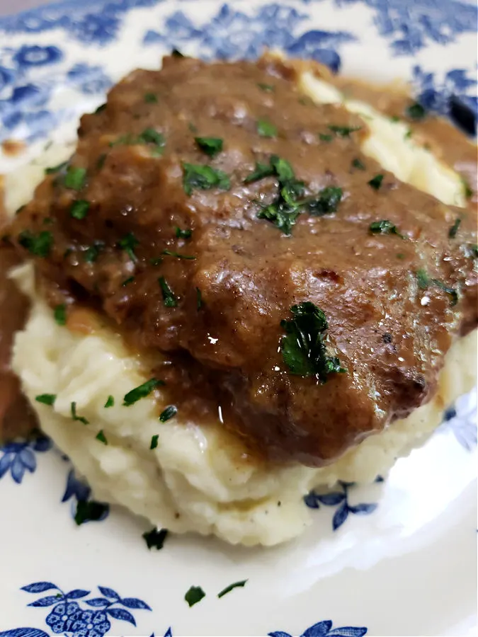 Serving of crock pot cubed steak and gravy on a bed of creamy mashed potatoes on a beautiful blue and white plate