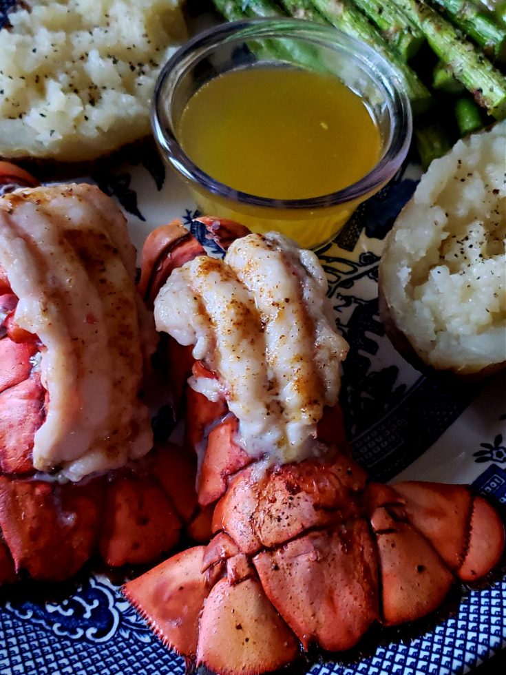 Broiled Lobster Tails on a serving platter with clarified butter, baked potatoes and asparagus