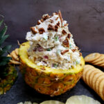 Easy Pineapple Cheese Ball Recipe with pecans, cheddar cheese, green pepper, onion, and seasoned salt served in a pineapple half with crackers