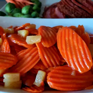 White serving bowl with glazed carrots with pineapple with a baked ham in the background