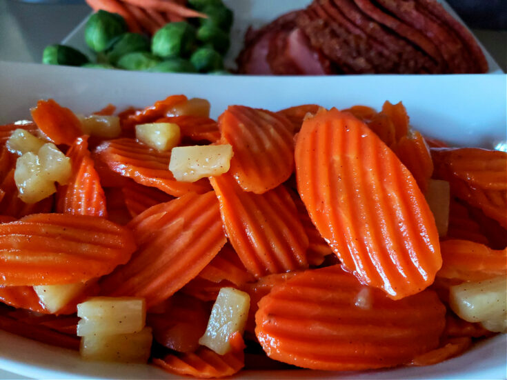 White serving bowl with glazed carrots with pineapple with a baked ham in the background