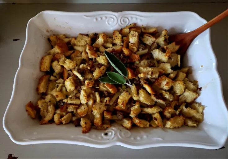 Old fashioned sausage stuffing, a Thanksgiving side dish, in a white baking dish with a wooden serving spoon. Garnished with fresh sage leaves