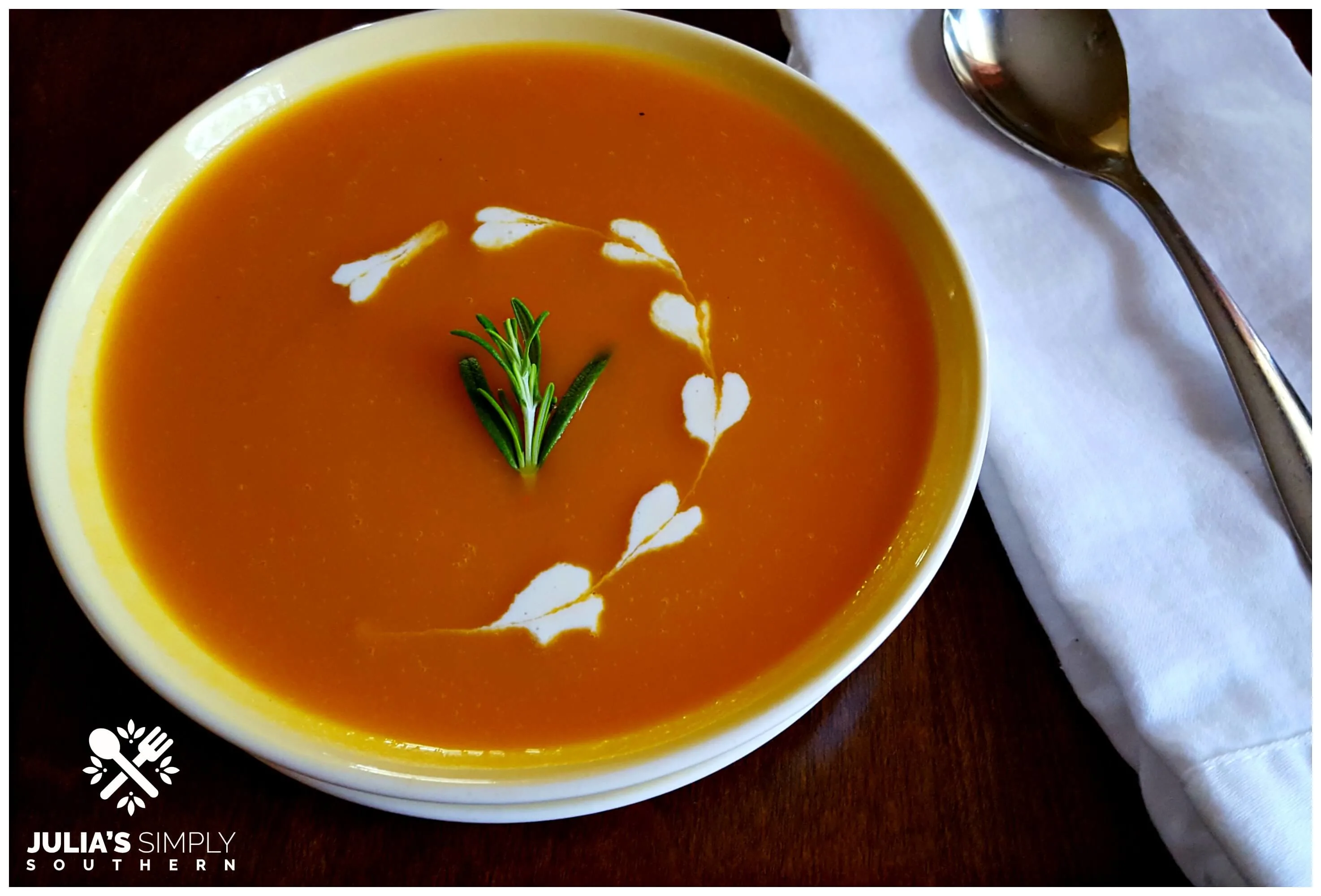 Bowl of the best pumpkin soup recipe with dots of heavy cream hearts and a sprig of Rosemary garnish