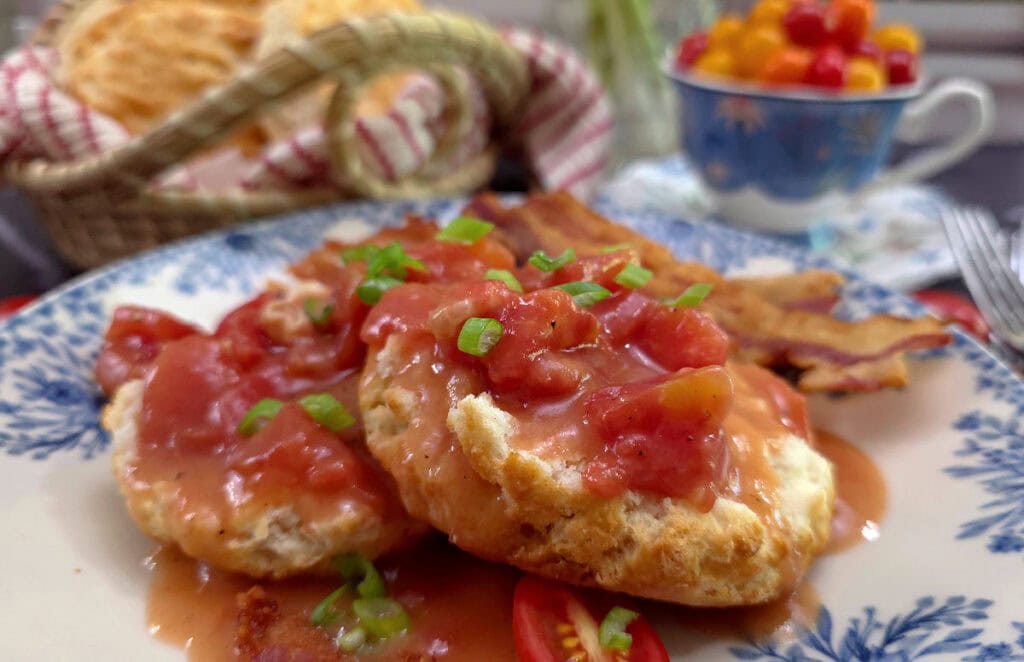 Old fashioned tomato gravy on a blue and white plate with cup of tomatoes in the background and a sweetgrass basket of biscuits