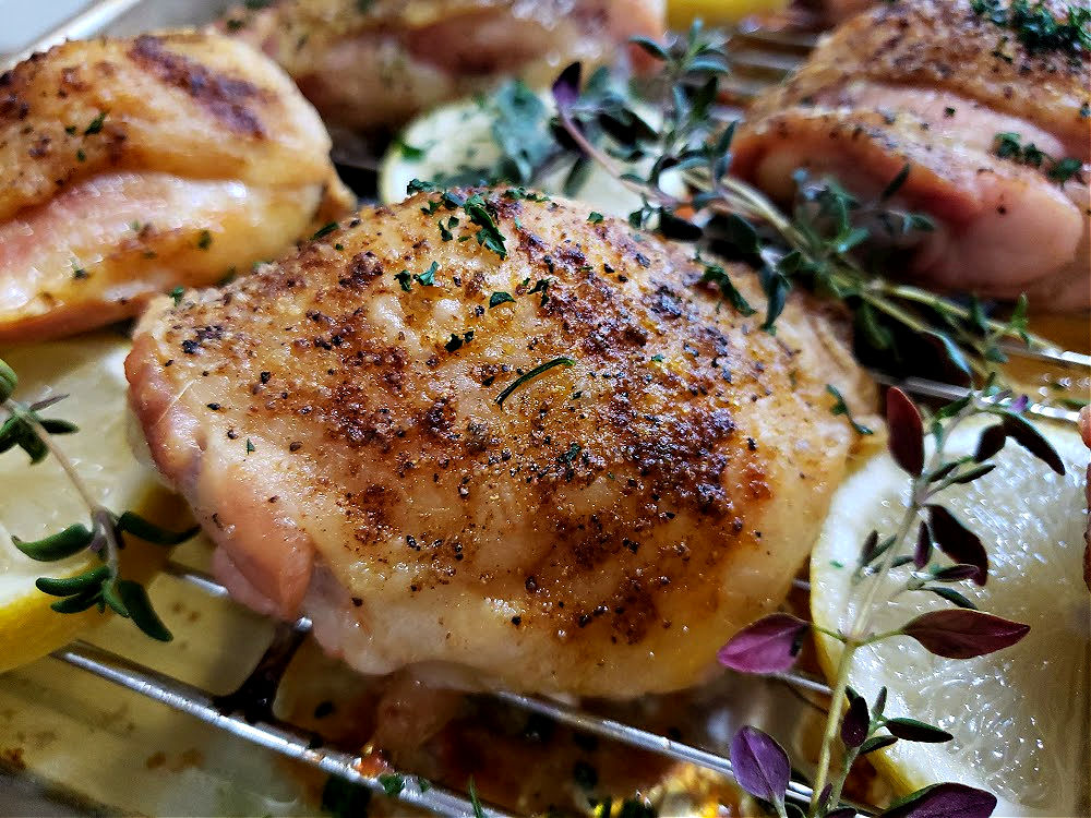 Oven Baked Chicken Thighs with Greek Seasoning. Crispy Skin and juicy on a baking rack pan garnished with lemon slices and fresh herbs.