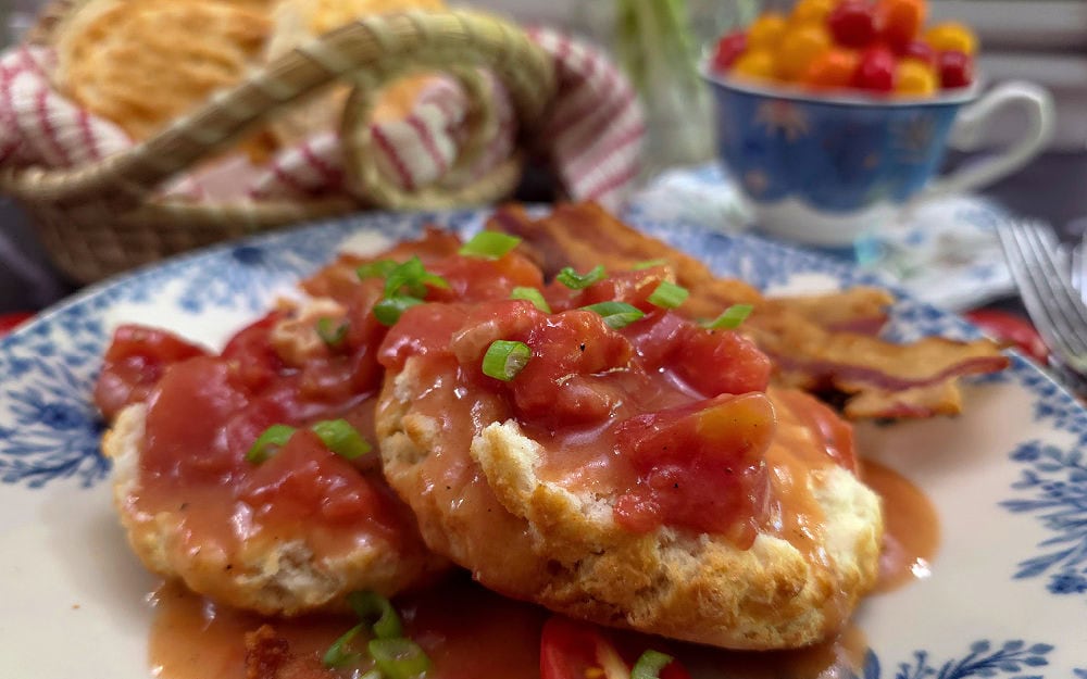 Blue and white plate with a biscuit topped with Southern tomato gravy with a sweetgrass basket full of biscuits in the background