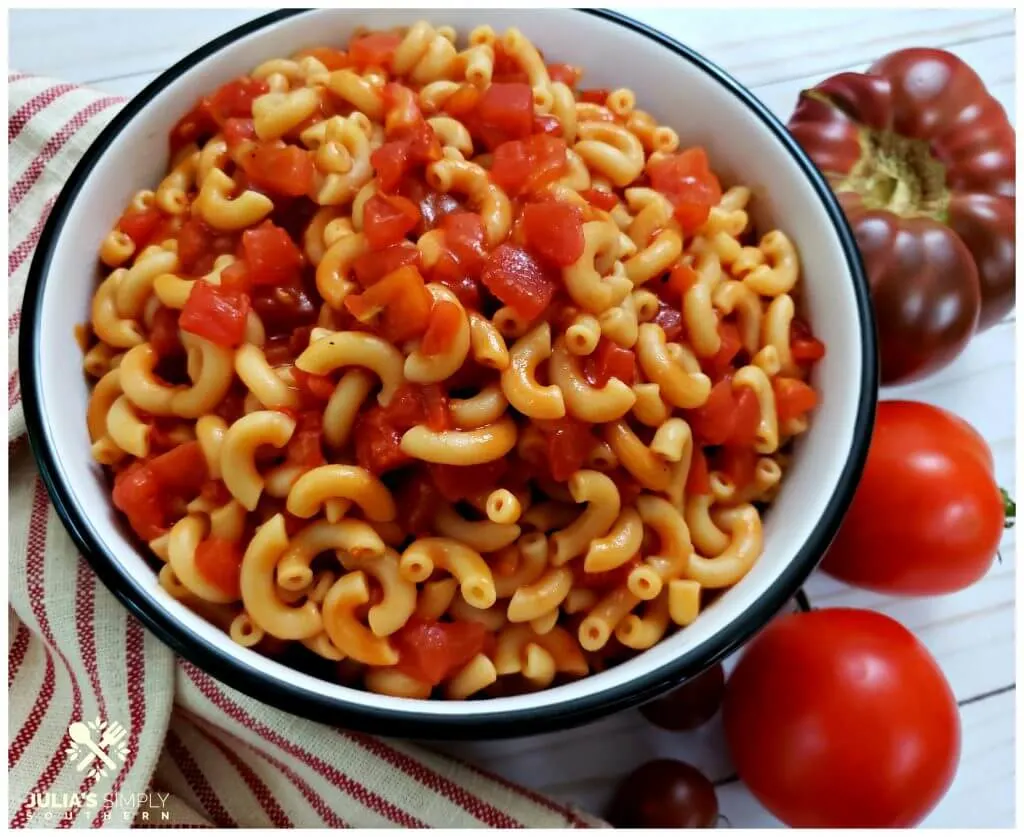 Bowl of classic macaroni with tomatoes surrounded by fresh garden tomatoes and a red ticking towel