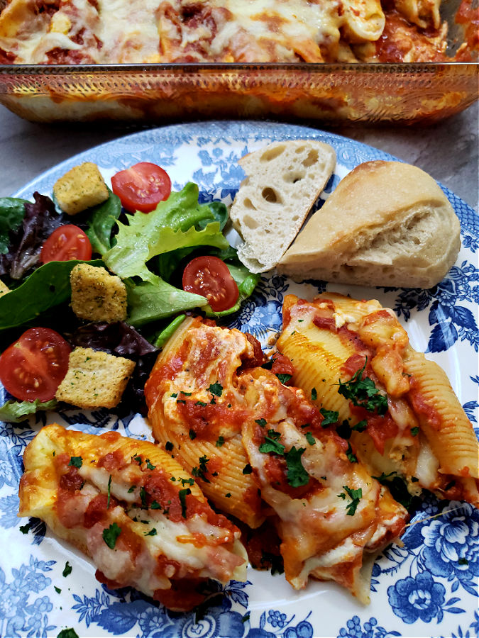 casserole dish with stuffed shells and a blue and white plate with stuffed shells with ricotta cheese filling with a simple salad and piece of Italian bread.