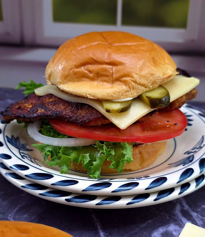 A plate with a blackened chicken sandwich with pickle, cheese, lettuce, tomato, onion on a toasted bun