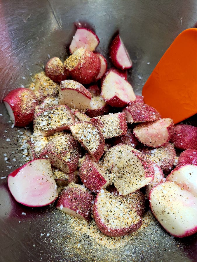 Stainless mixing bowl with radishes, olive oil, and seasonings being combined with a spatula prior to roasting in the oven