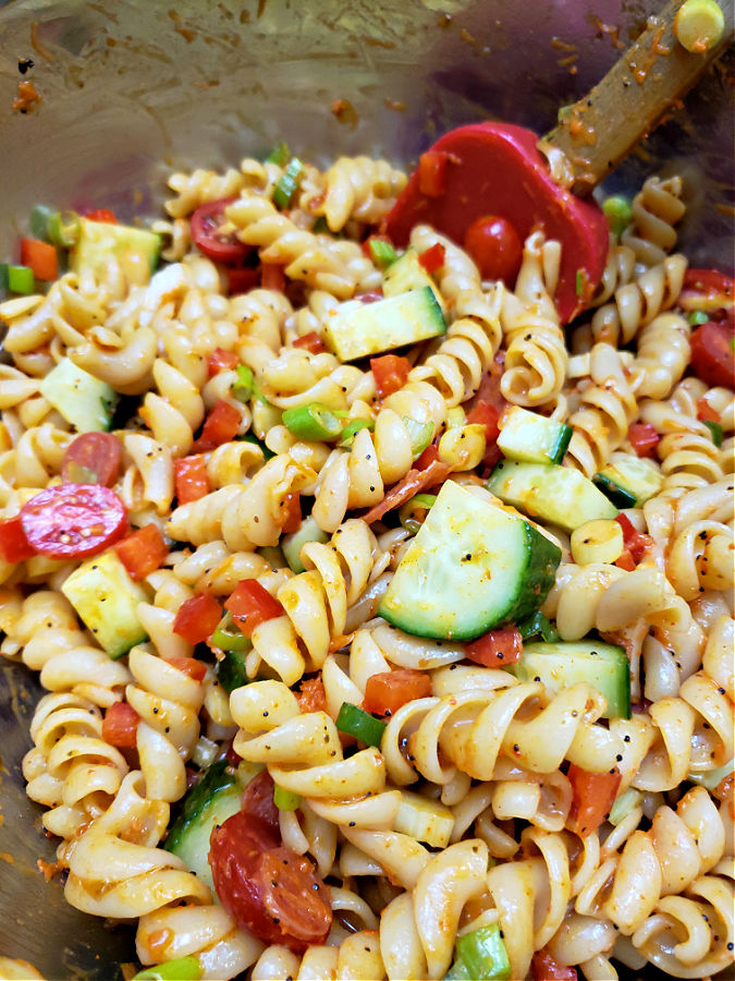 Using a spatula to stir together the pasta salad mixture in a large stainless mixing bowl