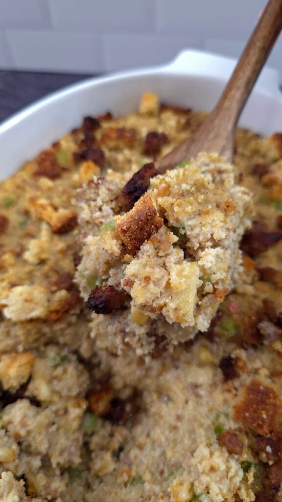 Wooden serving spoon serving close up of sausage and cornbread dressing with herbs and crispy topping.