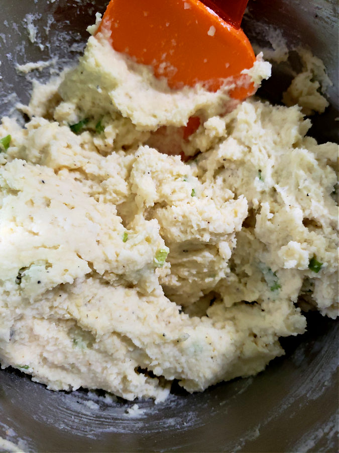 stainless mixing bowl with mixture combined for fried potato cakes made from leftover mashed potatoes being stirred with an orange spatula