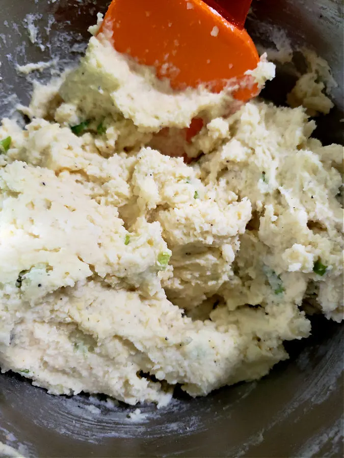 stainless mixing bowl with mixture combined for fried potato cakes made from leftover mashed potatoes being stirred with an orange spatula