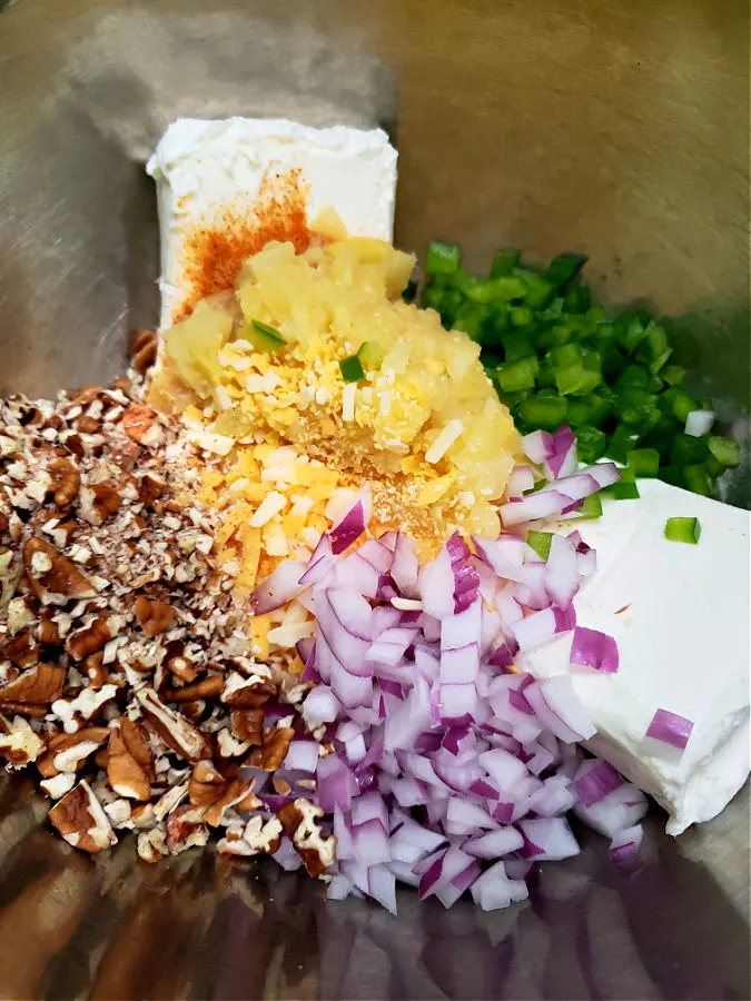 mixing bowl with ingredients for pineapple cheese ball recipe including cream cheese, crushed pineapple, red onion, green pepper, pecans, cheddar cheese, and seasoned salt
